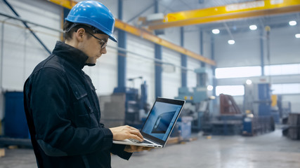 Factory worker in a hard hat is using a laptop computer with an engineering software.