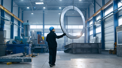 Factory worker in a hard hat is moving a metal detail with a remote crane.