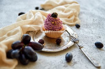 Purple golden cupcake with blackberry on silver vintage tray decorated with dessert fork, light brown cloth, branch of fresh blue grapes and blackberries on gray concrete background. Close up view