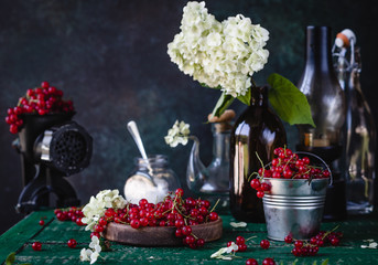 red currant berries in wooden bowl, metal bucket and in black hand meat chopper with white flowers, assorted bottles on a green wooden table