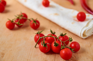 Branch of red cherry tomatoes isolated on light brown wooden background. On blurred background red chili pepper, tomatoes and pita. Side view