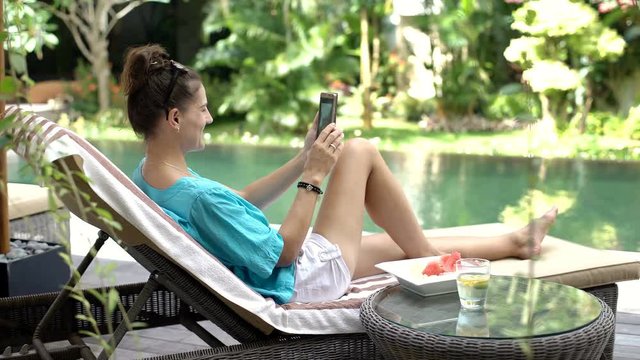 Happy woman sitting on sunbed next to the swimming pool and having a videocall on smartphone, steadycam shot
