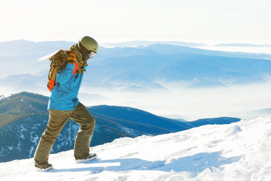 Young snowboarder in helmet walking to the very top of a mountain and carrying snowboard behind his back