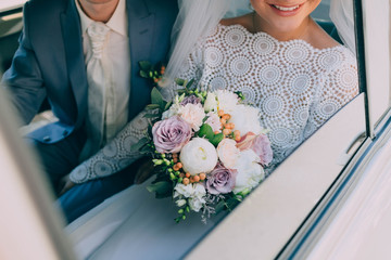 Beautiful bride in a white dress and groom in tuxedo are sitting in a car and holding a wedding bouquet. Groom embrace woman by the waist. Artwork - 168992487