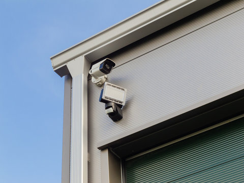 Nahaufnahme &Uuml;berwachungkamera und Au&szlig;enbeleuchtung mit Bewegungsmelder an einer Industriehalle - Close-up of surveillance camera and exterior lighting with motion detector on an industrial hall