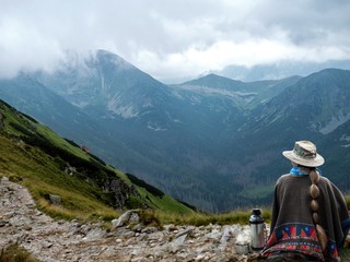 Fototapeta na wymiar Rest in the mountains. A woman in a poncho drinks tea from a thermos bottle.