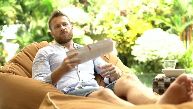 Man feels hot while sitting on the bean bag chair and cooling himself down with a gazette
