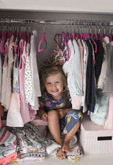 Beautiful little girl sitting in her wardrobe and smiling. Girl's clothes on hangers in the wardrobe.