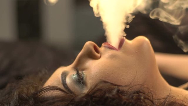 Glamour portrait of the woman smoking the hookah. She ies blowing out the smoke while laying on the bed. Side view.