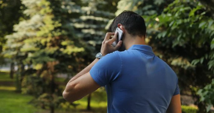A young attractive man in a blue T-shirt speaks by telephone in the park. Modern man. Green eyes. Clock on hand