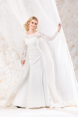 Fototapeta na wymiar fashionable gown, beautiful blonde model, bride hairstyle and makeup concept - smiling girl in wedding white dress standing indoors on light background, charming young woman posing near curtains