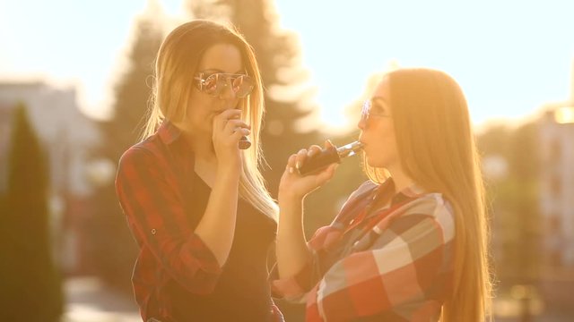 Two women vaping outdoor. The evening sunset over the city. Toned image.