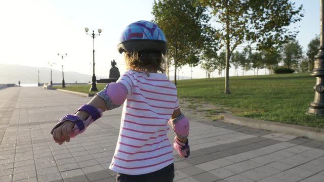 Little girl in helmet and protection riding on roller skates in summer park