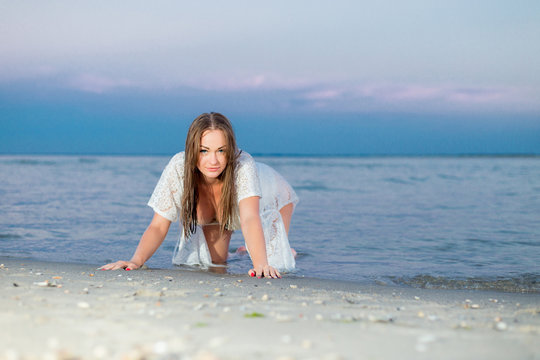 beautiful sexy girl in a white bathing suit and tunic sits on the shore of a deserted beach.