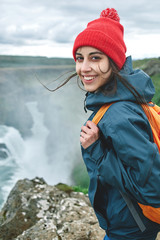 Girl in warm clothing, in red knitted hat and small orange backpack stands on the cliff on...