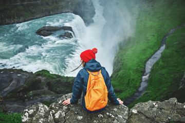 Girl in warm clothing, in red knitted hat and small orange backpack sits on the cliff on background of Gullfoss waterfall in Iceland. View from above