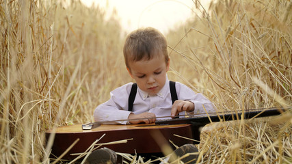 Naklejka na ściany i meble Little boy sitting in a golden wheat field and playing the guitar.