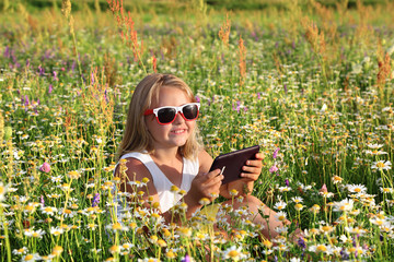 Śliczna uśmiechnięta dziewczynka bawi się tabletem wśród polnych kwiatów. - obrazy, fototapety, plakaty