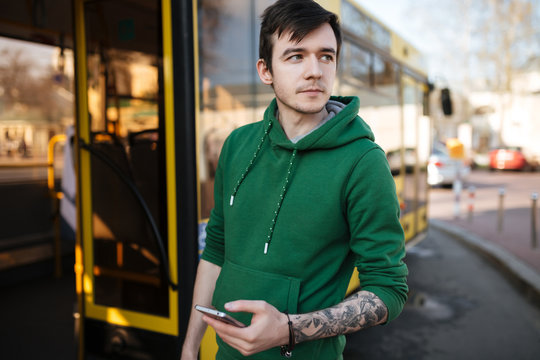Young man with dark hair standing outside with mobile phone in hand and bus on background. Portrait of thoughtful boy looking aside in green sweatshirt standing on street with cellphone