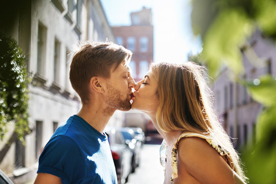 Profile outdoor portrait of handsome unshaven young male with fair hair giving a peck on lips of attractive woman. Beautiful sweet Caucasian couple in love kissing on street at the end of first date