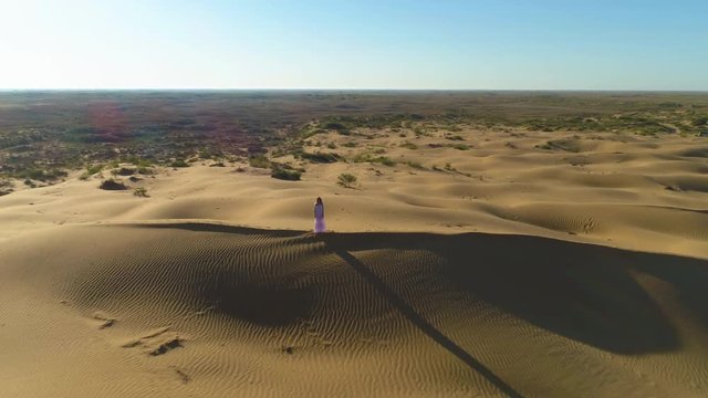 Drone is flying over young lonely woman with long hairs in white dress staying on the top of sand beige dune in desert. Aerial view.
