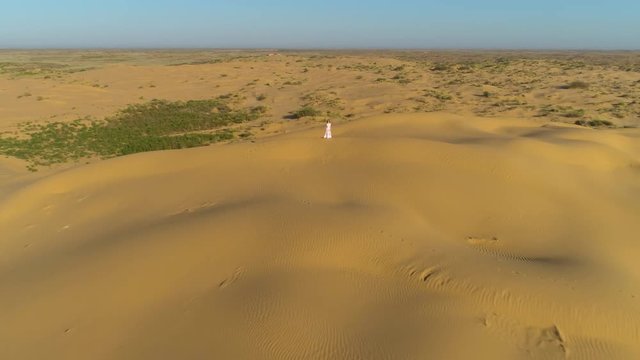 Young lonely woman with long hairs in white dress is staying on the top of sand beige dune in desert. Drone is distancing. Aerial view.