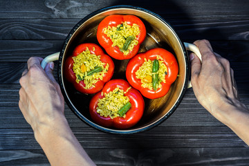 Chef holding a pan with four red stuffed peppers on dark rustic kitchen table background first person view. Cooking process top view