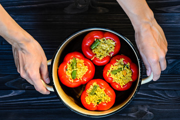 Chef holding a pan with four red stuffed peppers on dark rustic kitchen table background, top view. Cooking process