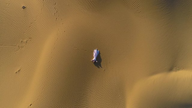 Young woman in white dress lies on golden sand in desert. Aerial vertical top shot. Drone is spinning around and flying upward..