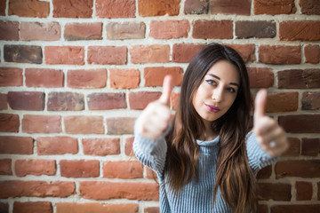 Beautiful young woman in casual wear with thumbs up, smiling, standing against white brick wall