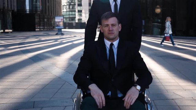 Young attractive businessman tote his cripple colleague in wheelchair outdoor, near business centre glass building. He looks sad or have no emotions on face. Slow motion sunrise shot.
