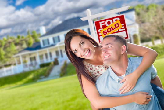 Playful Excited Military Couple In Front of Home with Sold Real Estate Sign.