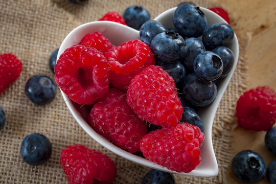 healthy foods and good eating habits concept with a heart shaped white bowl of blueberries and raspberries spilt in the middle, half blueberry half raspberry with berries and burlap in the background