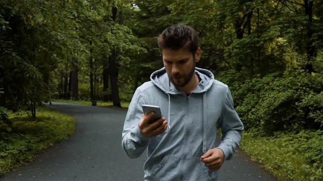 Young powerfull handsome runner in gray hook wear, jogging outside in autumn nature park with phone. Super slow motion shot with steadicam. Front middle footage