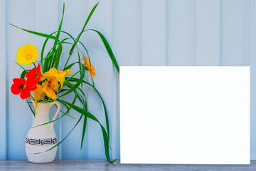 Mock up blank white paper sheet on light blue metal background with a vase with a bouquet of yellow flowers and green grass on a wooden table