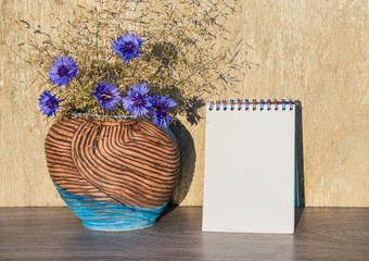 Mock up blank white sheet of paper on a wooden table with a vase and a bouquet of blue wildflowers and grass on background wooden wall vintage,