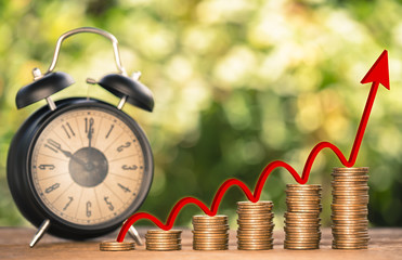 Money coin stack arranged as a graph on wood and black alarm clock with red arrow indicates money growth upturn and blur nature background, concept of time to money growth