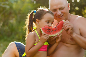 Grandfather and granddaughter eat watermelon and laugh in the garden on the grass in summertime