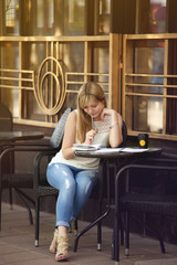 Pretty blonde girl copywriter sitting on cafe terrace with coffee. Woman freelancer writing outdoors.