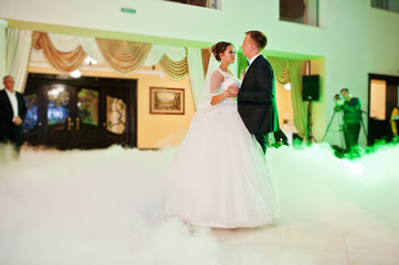 Fototapeta premium Beautiful wedding couple dancing their first dance in the huge hall with heavy smoke, different lights and people looking at them.