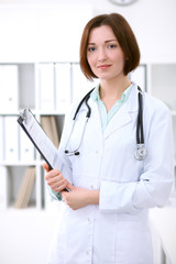 Young brunette female doctor standing with clipboard and smiling at hospital.