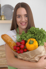 Young caucasian woman in a green apron is holding paper bag full of vegetables and fruits while smiling in kitchen