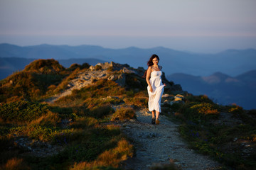 woman running outdoor in mountains at summer sunset. Smiling happy woman. Young woman in long white dress. Young woman running on mountain peak. Girl running outdoor in mountains at summer sunset