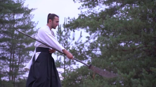 A warrior in a kimono with a tattoo on his shoulder emerges from the forest and becomes in fighting posture with a halberd