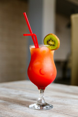 A colourful, bright red and orange cocktail in a high glass with a slice of green kiwi and two red...