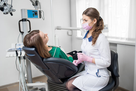 Young female dentist with beautiful woman patient before dental procedure in modern dental office. Doctor wearing white uniform, pink gloves and mask, holding dental tools. Dentistry