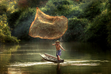 Thai fisherman on wooden boat casting a net for catching freshwater fish in nature river in the...