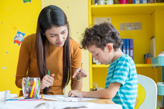 Asian woman teacher teaching boy kid to paint color book on table in classroom,kindergarten education school