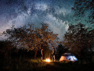 Man and woman hikers standing at a campfire near tent under trees and beautiful night sky full of stars and milky way. Night camping. Astrophotography