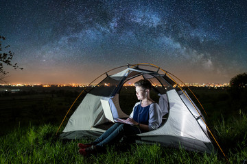 Woman using her laptop with headset in the camping at night. Female sitting in the tent under beautiful starry sky and milky way. On the background luminous city. Astrophotography